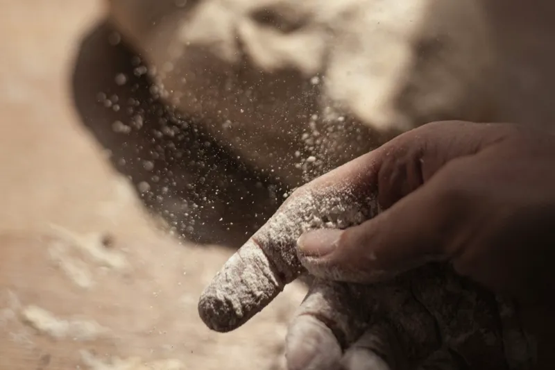 A floured hand over the dough, flour falling onto the wooden board